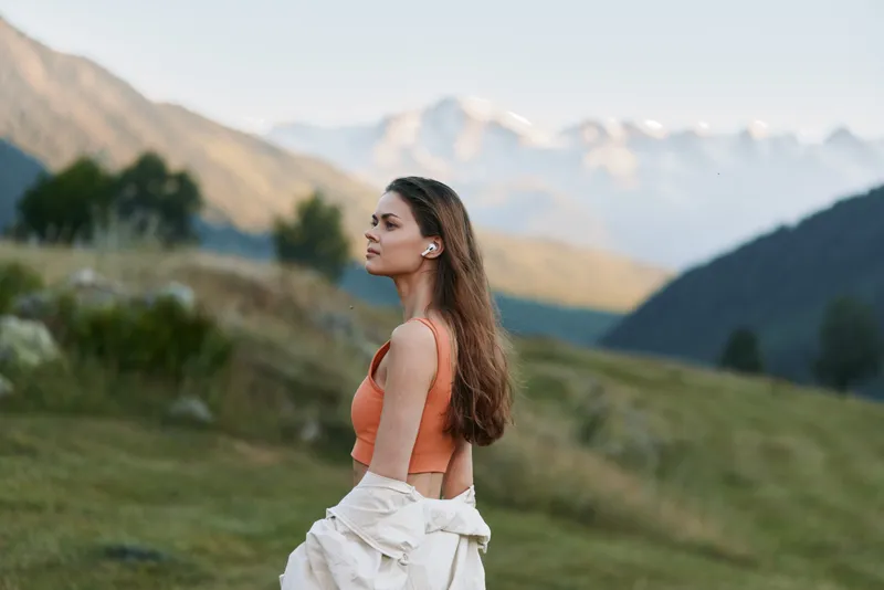 A young woman stands in a mountainous landscape, wearing an orange top and a light jacket over her shoulders. She gazes into the distance, calm and contemplative in nature.