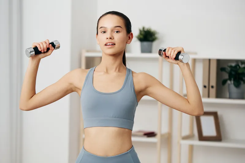 Female fitness enthusiast raises dumbbells in a bright home gym, performing a shoulder raise with strong posture. Clean shelves, plants and a neat workout space frame dedication.