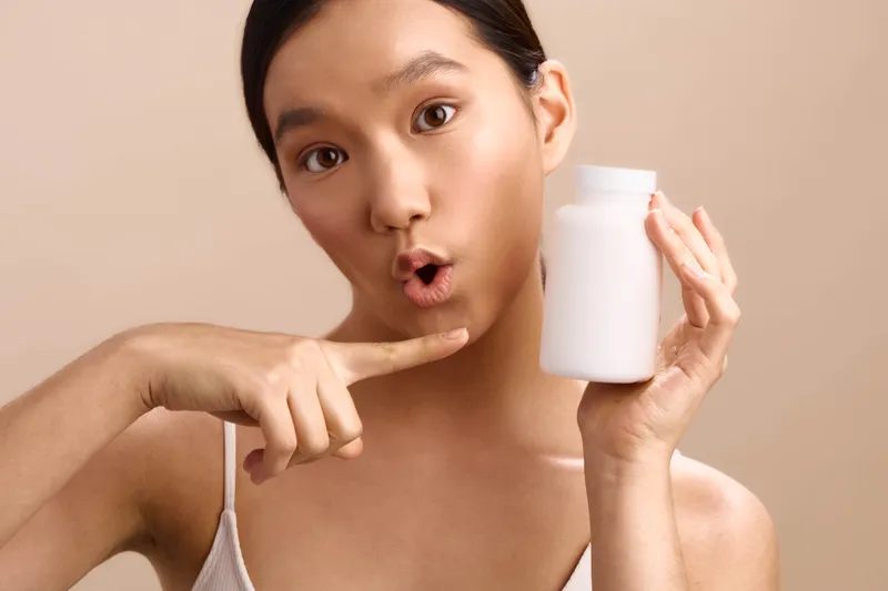 asian woman holds a white bottle close to her face, eyes wide and lips puckered, pointing to the bottle with a curious expression, in a clean studio environment suggesting wellness.