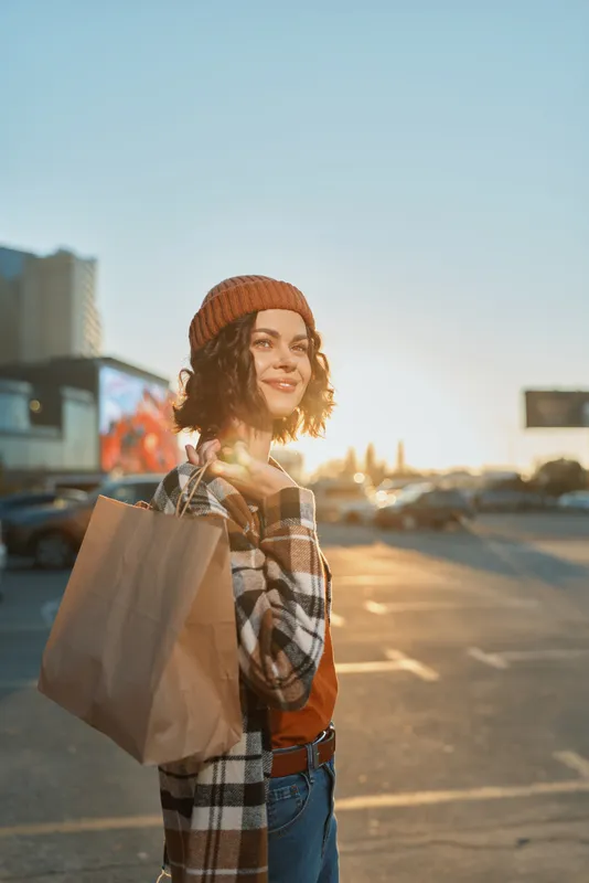Woman shopping portrait in urban parking, golden hour glow lighting her beanie and smile. Candid lifestyle scene with authenticity, mindful living and emotional storytelling moment.
