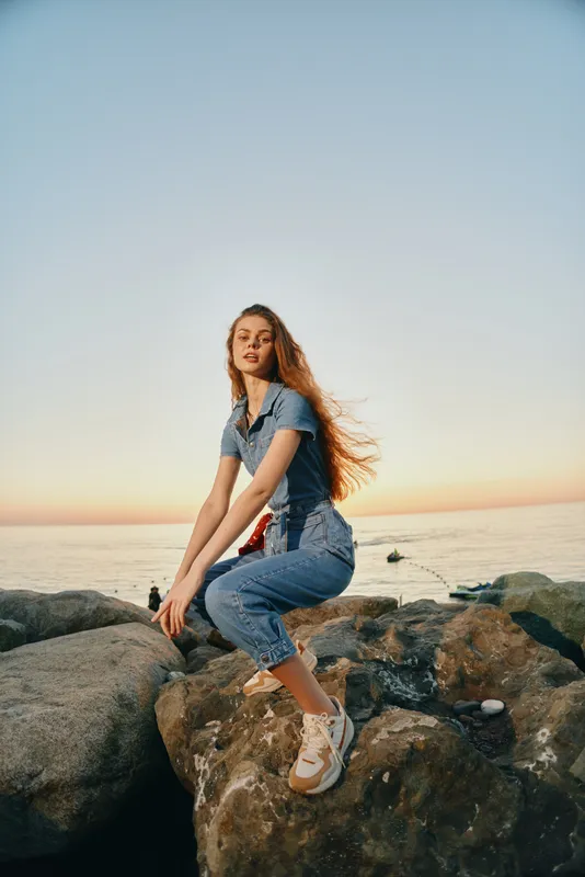 lifestyle woman in modern boho-western denim street style sits on rocks by the sea at sunset with film color tones and natural light enhancing the mood