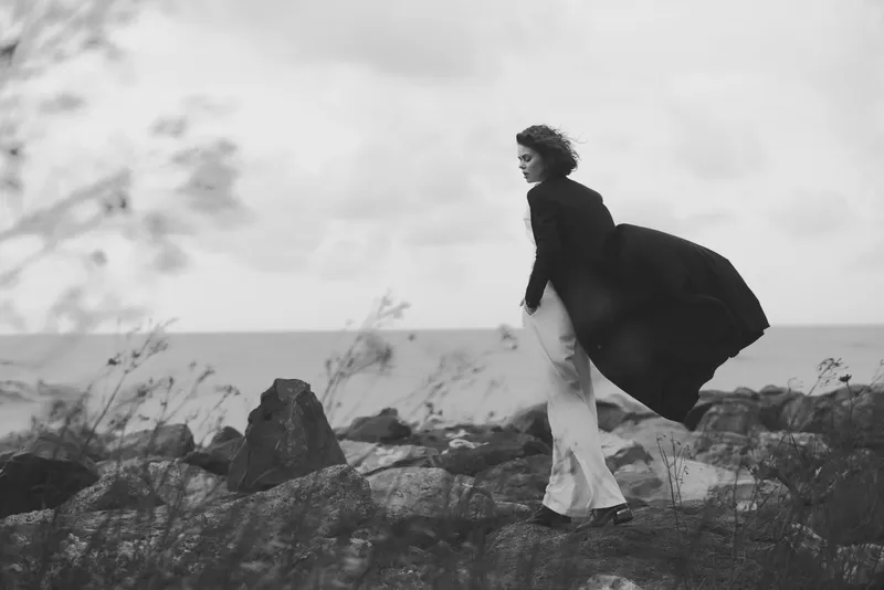 Woman walking along a windy coast, cape fluttering behind, amid rocky terrain and sparse grass, black and white scene capturing movement, solitude, and a sense of journey against the seascape.