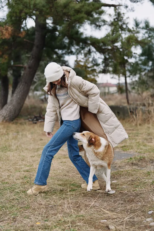 Person in a warm coat and beanie plays with a fluffy dog in a park, outdoor scene with trees, casual clothing and playful interaction in natural light