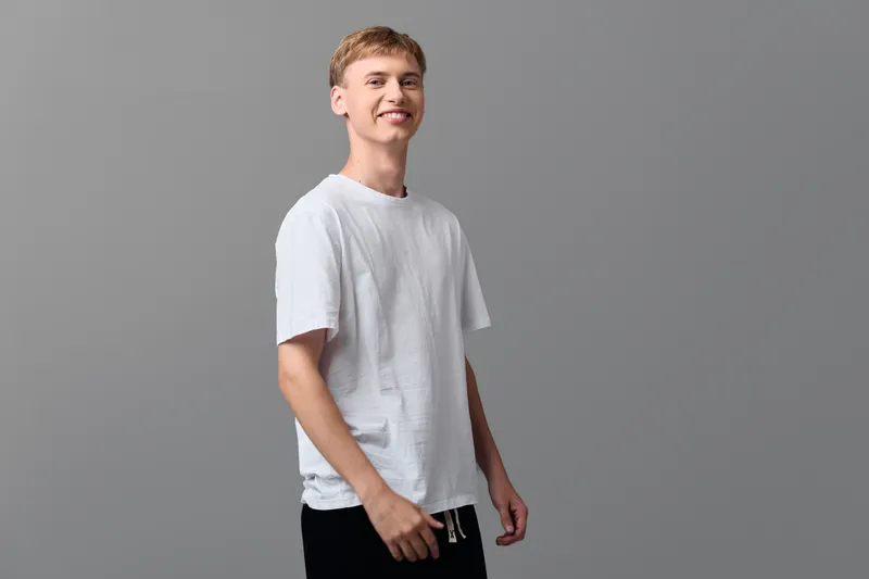 Casual young man in a white tee against a gray studio backdrop, standing with a relaxed pose and subtle smile, suitable for lifestyle fashion and everyday style themes