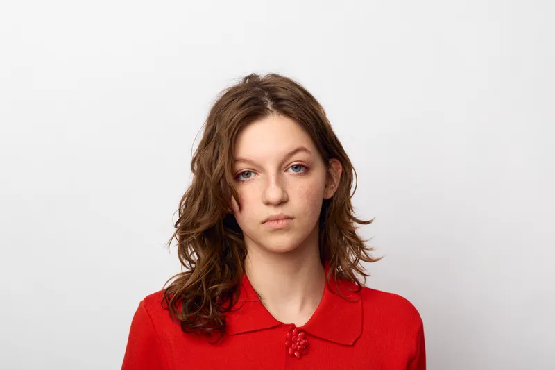 Young woman with curly brown hair wearing red sweater looks serious and thoughtful against white background. Concept of contemplation, emotion, and natural beauty portrait.