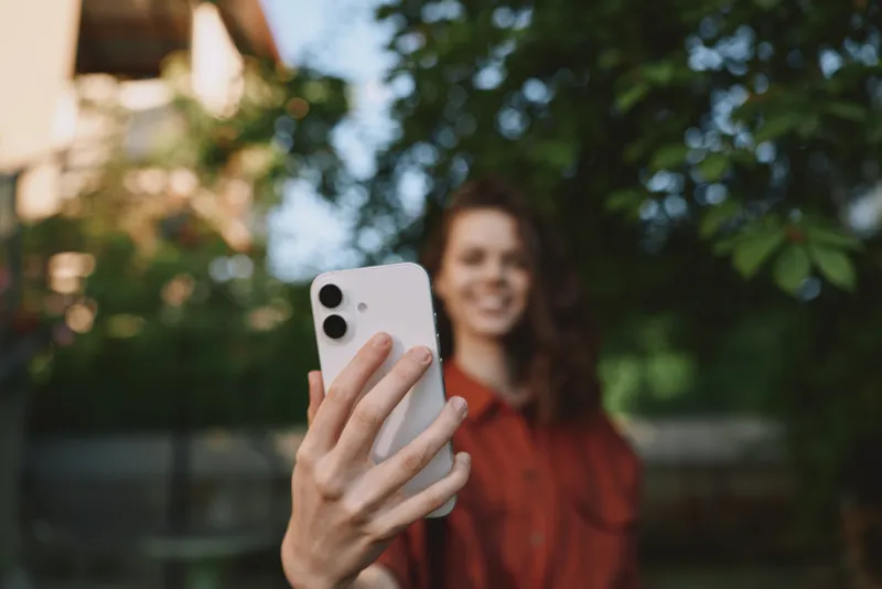 Smiling woman holding a phone for a selfie outdoors, soft natural light, casual lifestyle moment, blurred green background, vibrant mood, urban park vibe