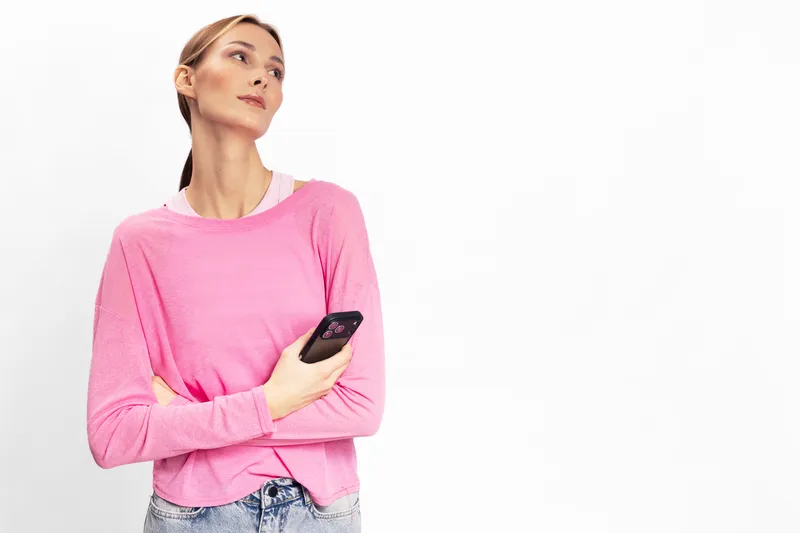 Young woman in pink sweater holding smartphone looks away thoughtfully on white background. Concept for communication, technology, lifestyle, and contemplation.