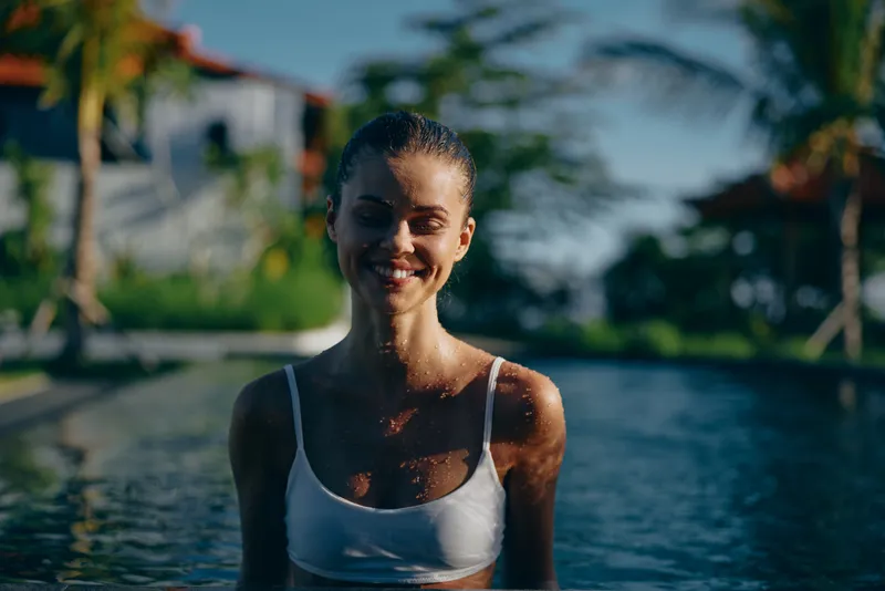 Young woman in white swimsuit stands in outdoor pool smiling with closed eyes enjoying sunny day surrounded by tropical plants and blue sky background.