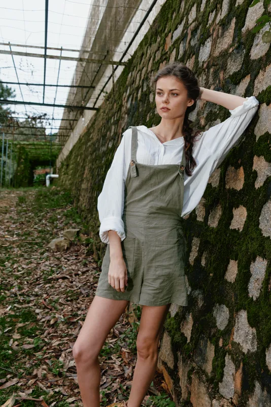 Young woman in stylish green overalls and white shirt stands against a rustic stone wall, showcasing a relaxed, natural fashion style perfect for summer