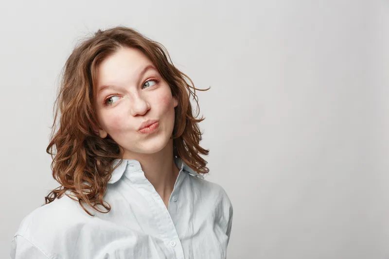 Young woman with curly hair wearing white shirt looks sideways with playful expression on light background. Concept of casual style, youth, and natural beauty.