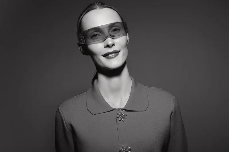 Elegant white woman with pale skin wearing modern glasses and vintage textured jacket against studio background.