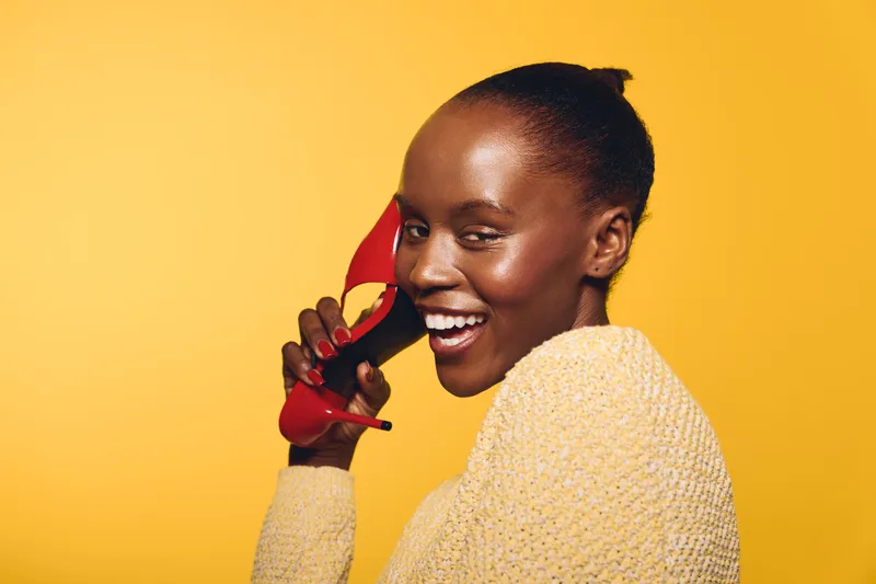 Smiling dark skinned African woman with short hair holds red vintage phone receiver near ear. Yellow background and cozy knitted sweater create warm and cheerful communication concept.