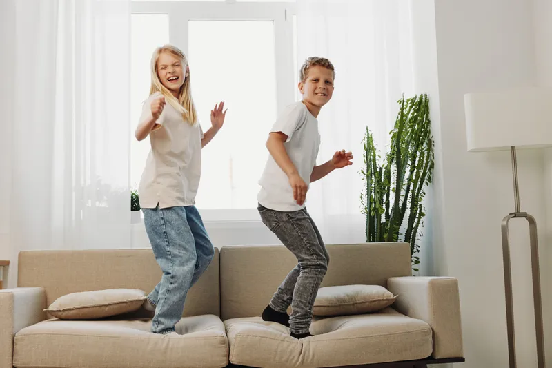 Two cheerful children jump on a beige sofa in a bright living room, enjoying playful moments and carefree indoor fun together. Natural light fills the space.