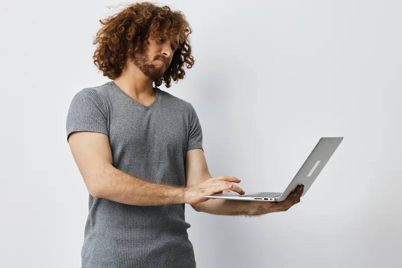 Young man with curly hair using a laptop, wearing casual gray shirt, focused on work, minimalist background, ideal for business and technology themes