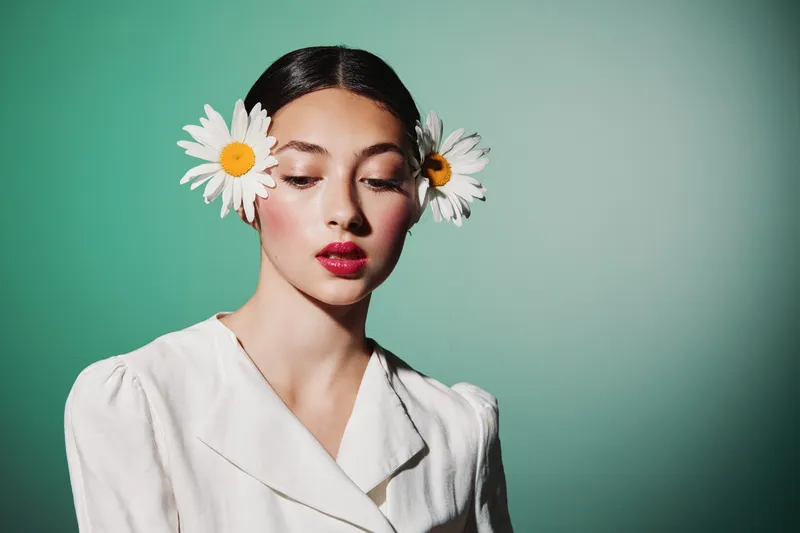 Young woman with bright red lipstick wearing white vintage outfit and daisy flowers in hair on green background. Concept of beauty, fashion, creativity.