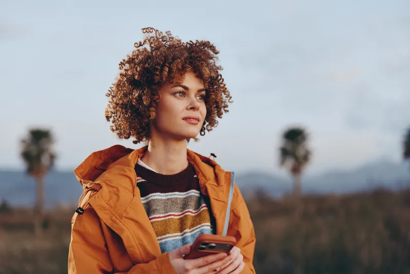 Woman lifestyle, curly hair, outdoor portrait of a woman in orange jacket holding a smartphone, sunny landscape with soft mountains, warm evening light, modern communication concept
