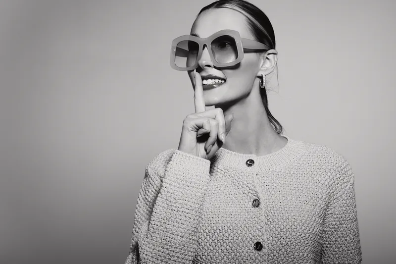 Stylish Caucasian woman with light skin wearing large sunglasses and a knitted sweater holds finger to lips in shushing gesture. Concept of secrecy, fashion, and elegance in studio.