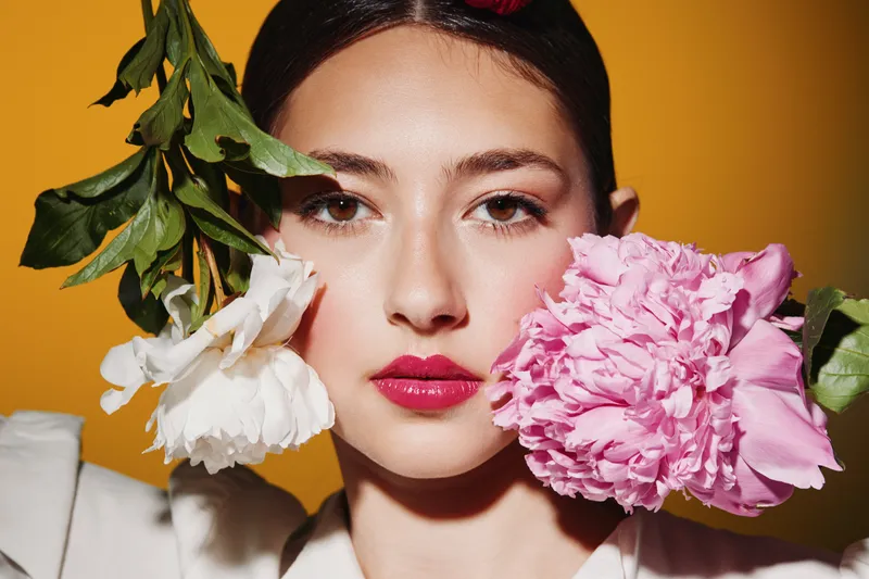 Close-up of a young woman with glossy pink lips and dewy skin creatively posed with vibrant pink and white flowers at her cheeks. Bold beauty and fashion concept.