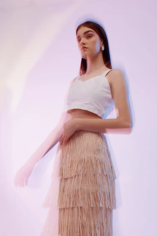 Young woman in a white crop top and beige fringe skirt poses against a soft pastel background. The image highlights beauty, fashion, and elegance with a dreamy atmosphere.