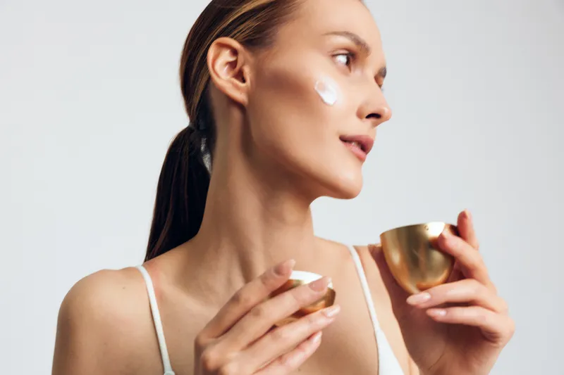Woman holding tea cup, skincare cream on face, close up profile, facial spa wellness beauty hydration, moisturizing routine, relaxing calm mood, clean gray background, golden cup
