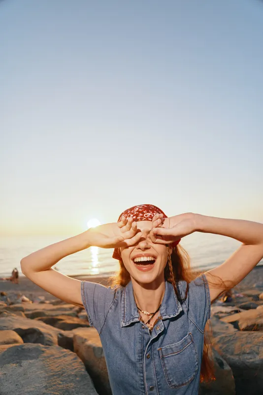 Lifestyle woman wearing modern boho-western denim street style laughing joyfully at sunset by the rocky shore with warm film color tones creating a relaxed vibe.