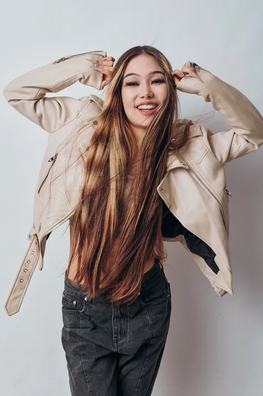 Young woman smiling with long hair and beige jacket, posing happily against light background. Casual style with natural expression and relaxed confident posture in studio.