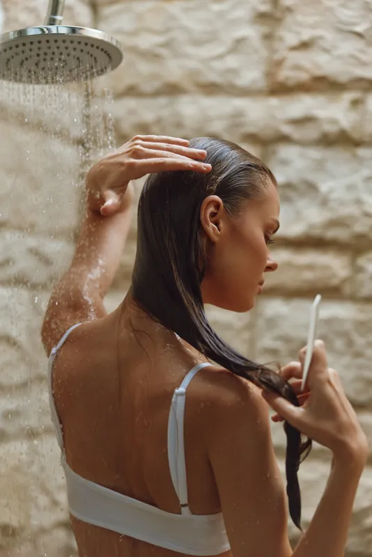 woman showering in natural light with wet hair under rainfall showerhead, serene mood, skin care routine, warm beige background texture