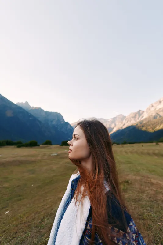 Woman profile portrait in a meadow with mountains and nature landscape, young female outdoors at golden hour. Longhair sideview, thoughtful serene expression and panoramic scenery.