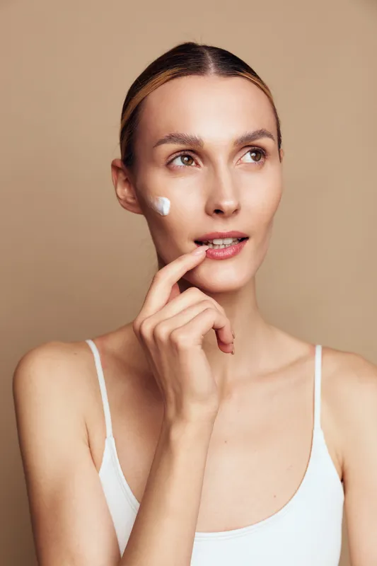 Young woman with clean skin applies face cream on cheek, looks thoughtful, wearing white tank top, isolated on beige background, natural skincare and beauty concept.