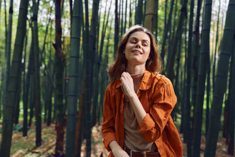 Woman smile portrait in bamboo forest, nature outdoors scene with casual orange jacket, relaxed happy adult exploring tall green trees, sunlight and serene mood