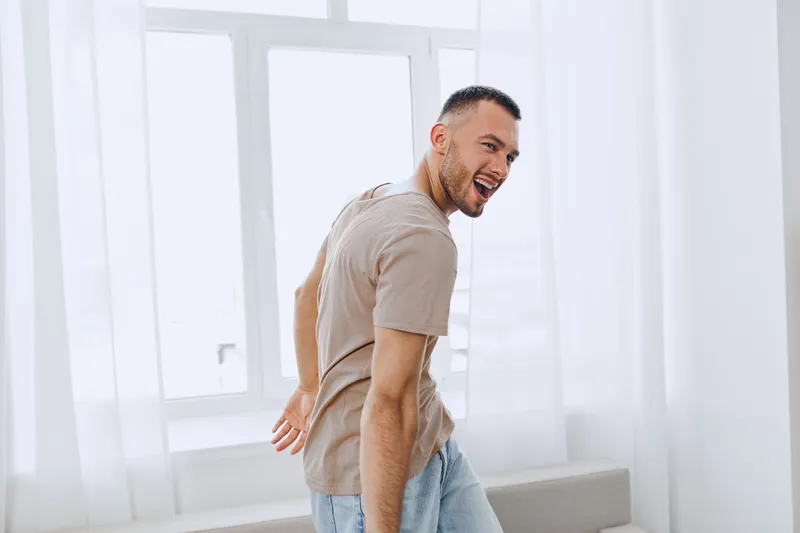 Joyful man smiling while dancing near a window, showcasing a vibrant atmosphere with bright, soft sunlight illuminating his carefree spirit.
