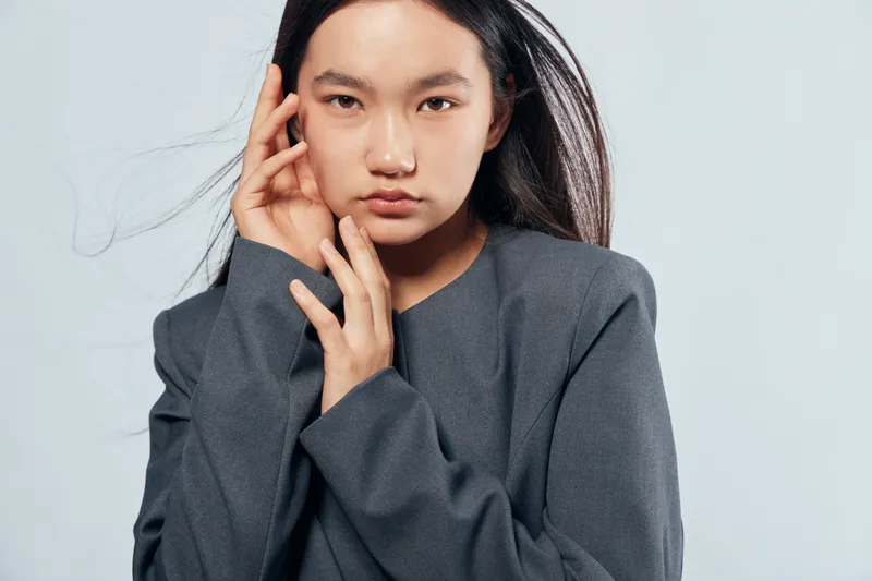 Young woman with long black hair and serious expression wearing dark gray sweater posing with hand near face on light background in studio portrait for fashion and beauty.