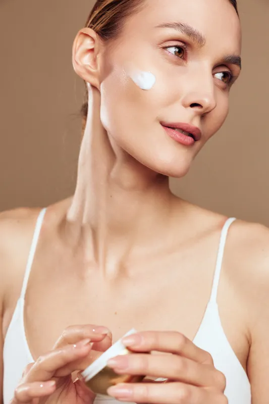 A young woman in a white tank top applies cream to her clean face against a neutral background, demonstrating skincare routine and natural beauty care concept.