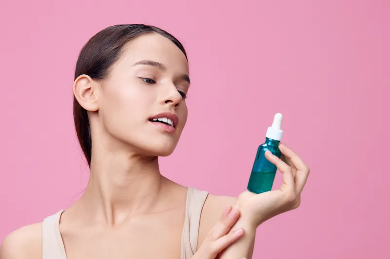 Young woman showcasing a blue serum bottle against a pink background, reflecting confidence and beauty, perfect for skincare and wellness themes