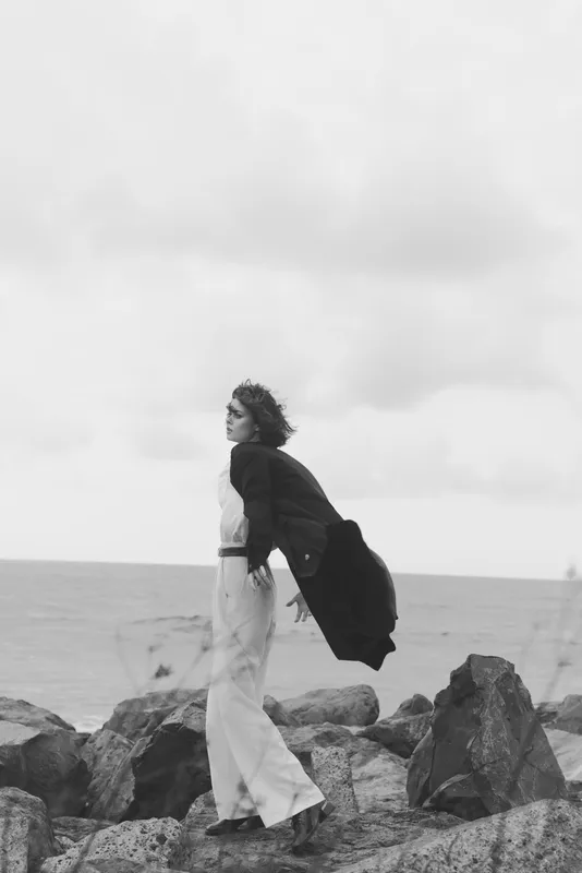 Woman walking along a rocky coast, wind catching her coat in a black and white seascape, capturing movement, atmosphere, and outdoor photography with dramatic sky and open horizon