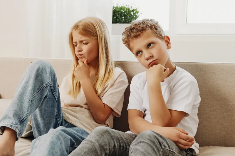 Thoughtful children sit on a sofa indoors, both resting chins on hands and looking away. The boy and girl appear pensive and contemplative in casual clothes near a bright window.