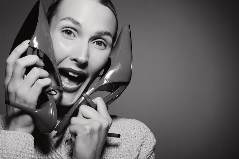 Young white woman with light skin holds shiny high heel shoes near face with joyful expression on grey background. Concept of fashion, style, and happiness.