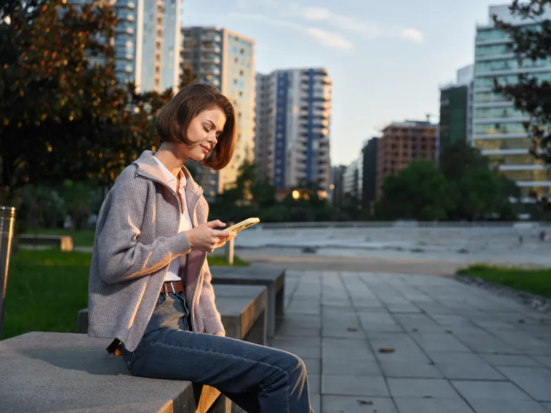 A cheerful woman sits on a concrete bench in a city park, absorbed in her smartphone, with warm sunset light and tall modern buildings lining the background.
