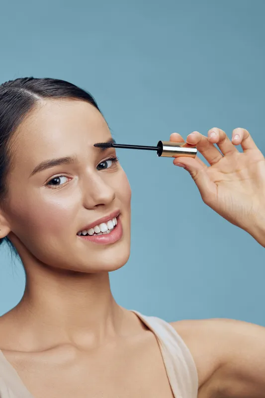 Young woman applying eyebrow gel, smiling confidently against a soft blue background, showcasing beauty and self care in a fresh, modern style