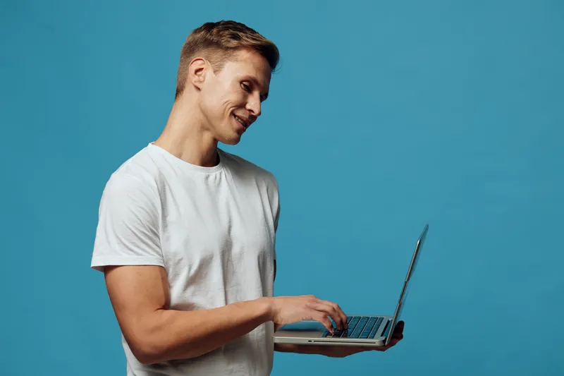 Young smiling man with light skin wearing casual white t-shirt hold laptop computer in hands look at camera isolated on plain solid blue background studio portrait. People lifestyle concept