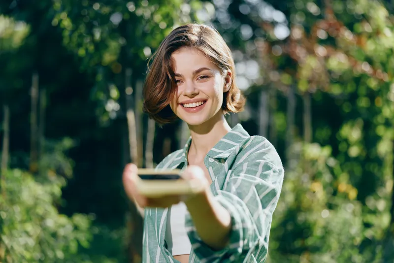 woman, outdoor, portrait, smile a cheerful woman holds a smartphone toward the camera in a sunny park, radiating happiness and connection with nature and the moment