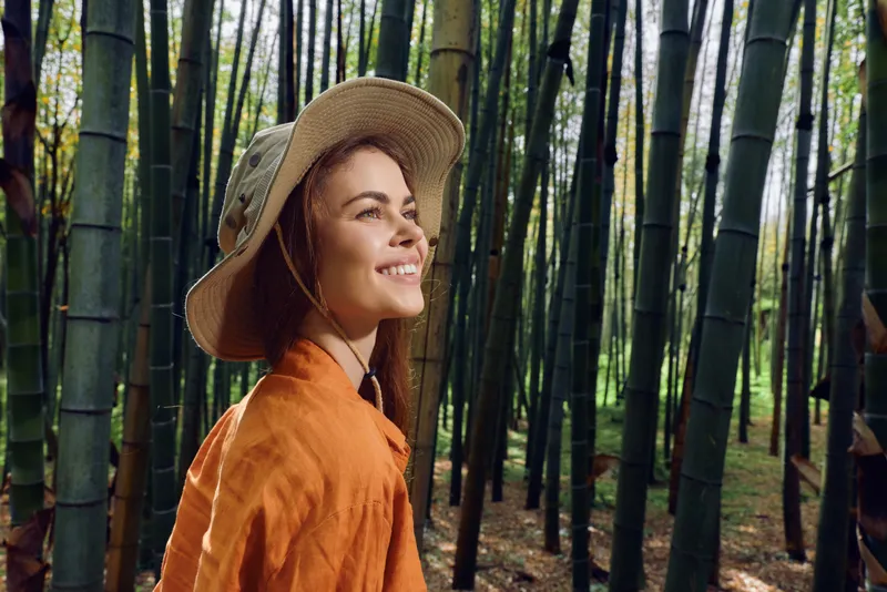 Woman in straw hat smiling in a bamboo forest portrait, nature travel scene with warm sunlight, orange jacket and peaceful mood, outdoor adventure and eco tourism.
