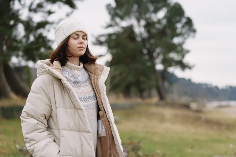 Woman wearing a thick winter coat and beanie stands outdoors in a breezy park, calm expression, soft natural light, highlighting cozy outerwear and a serene seasonal mood.