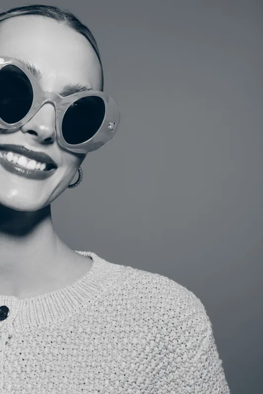 Smiling young white woman with pale skin wearing large round sunglasses and knitted sweater. Close-up portrait with natural makeup on gray background.