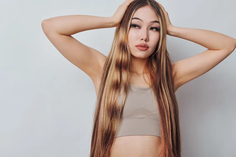 Young woman with long straight hair and natural makeup posing with hands on head against light background, wearing casual sleeveless top for beauty and fashion concept.