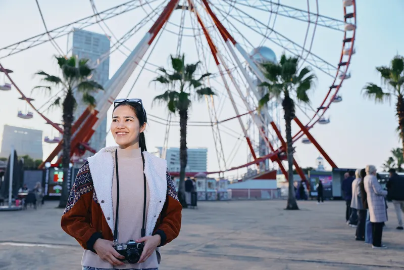 Smiling woman with camera at an outdoor amusement park beside a towering ferris wheel, capturing a lively moment of leisure, travel, and urban recreation under bright daylight