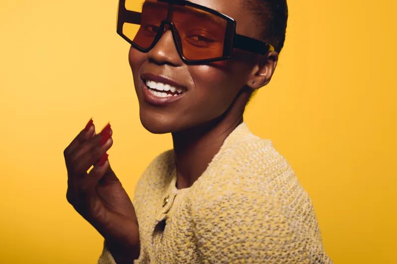 Stylish smiling black woman with dark skin wearing large sunglasses and a knitted sweater poses against yellow background. Concept of fashion, beauty, and confident African ethnicity.