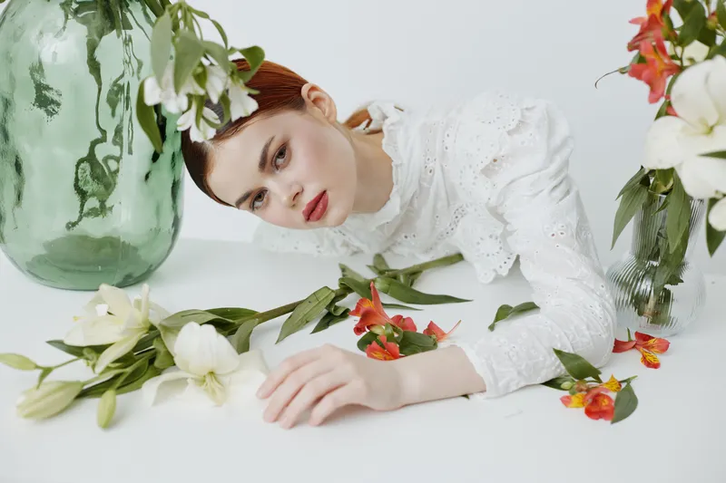 Elegant young woman in white lace blouse surrounded by green vase and fresh flowers, conveying a serene and natural beauty concept