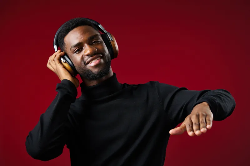 Happy young Black man enjoying music with headphones against a vibrant red background, showcasing joy and rhythm in a stylish black turtleneck