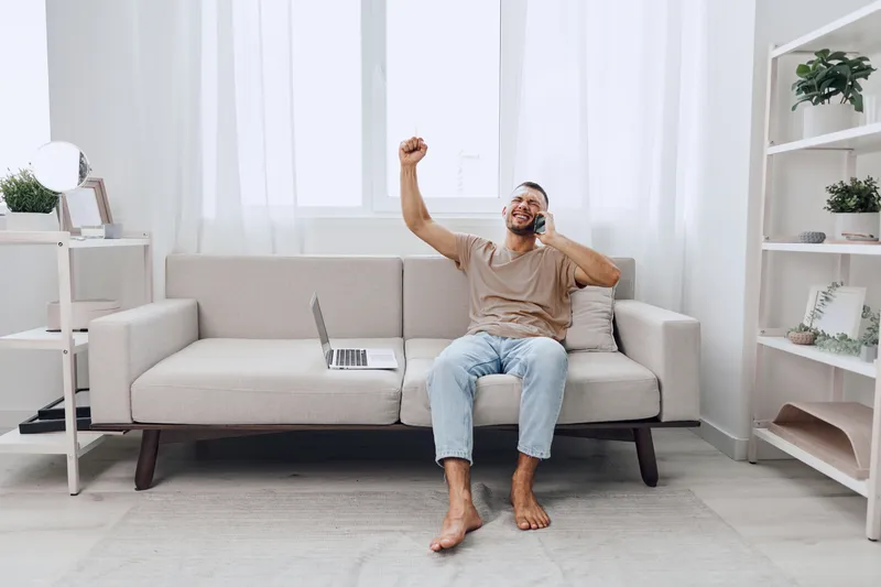 Happy man celebrating a successful work achievement while talking on the phone, dressed casually in a cozy home environment, showcasing modern lifestyle and comfort.